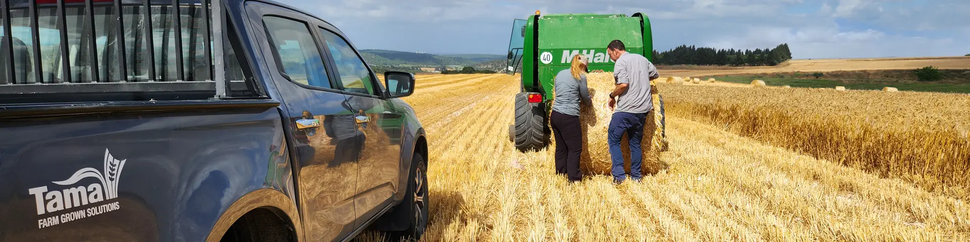 Tama field team inspecting a round straw bale wrapped with netwrap beside a McHale round baler in a harvested wheat field, with Tama Farm Grown Solutions service vehicle in the foreground.