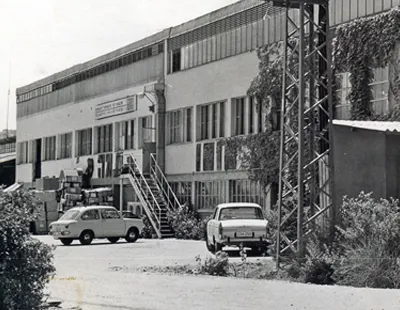 Black and white historic photo of Tama’s original factory building in Kibbutz Mishmar HaEmek, Israel, with vintage cars parked outside and early industrial facilities in the background.